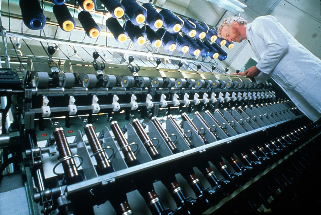 A technician in a white coat leans over a long spinning frame with rows of spindles and bobbins of wool yarn