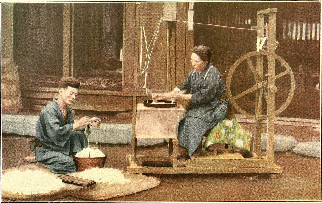 Hand-coloured early photograph of a woman reeling silk at a wooden wheel while a man sorts cocoons on the floor