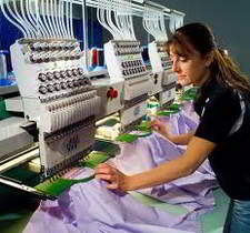 An operator positions a hooped garment under one head of a multi-head embroidery machine; each head carries a row of needles threaded with different colors